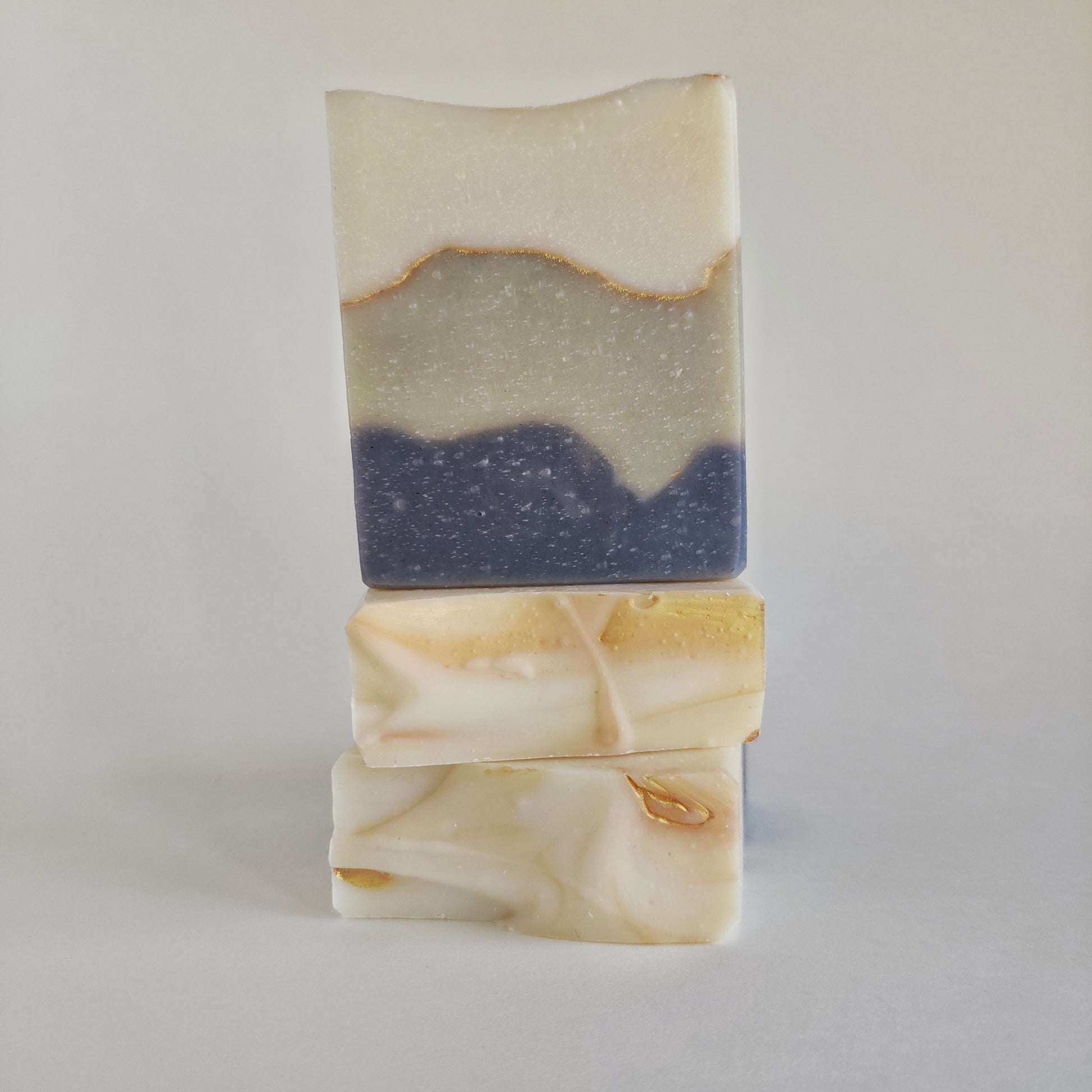 Product photo of three bars of soap from the front and top. The soap is decorated with white, green, and blue layers throughout. The top is decorated with additional gold swirls.